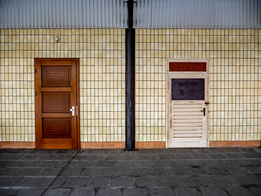 A photograph of a building's external wall, with pale yellow tiles. There are two doors, the left is brown wood and the right is painted white.