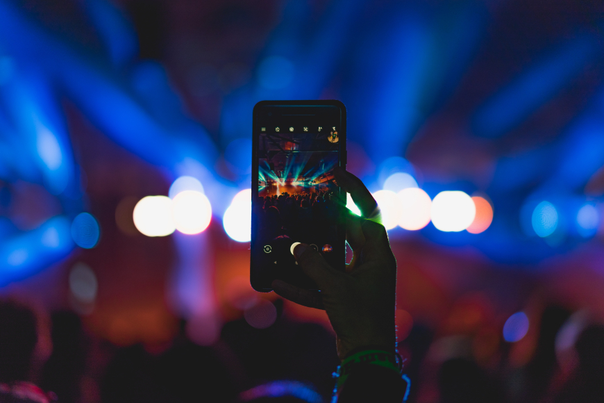 The silhouette of a hand holding up a smartphone and taking a photo or video of a live event with pixellated lights in the distance.