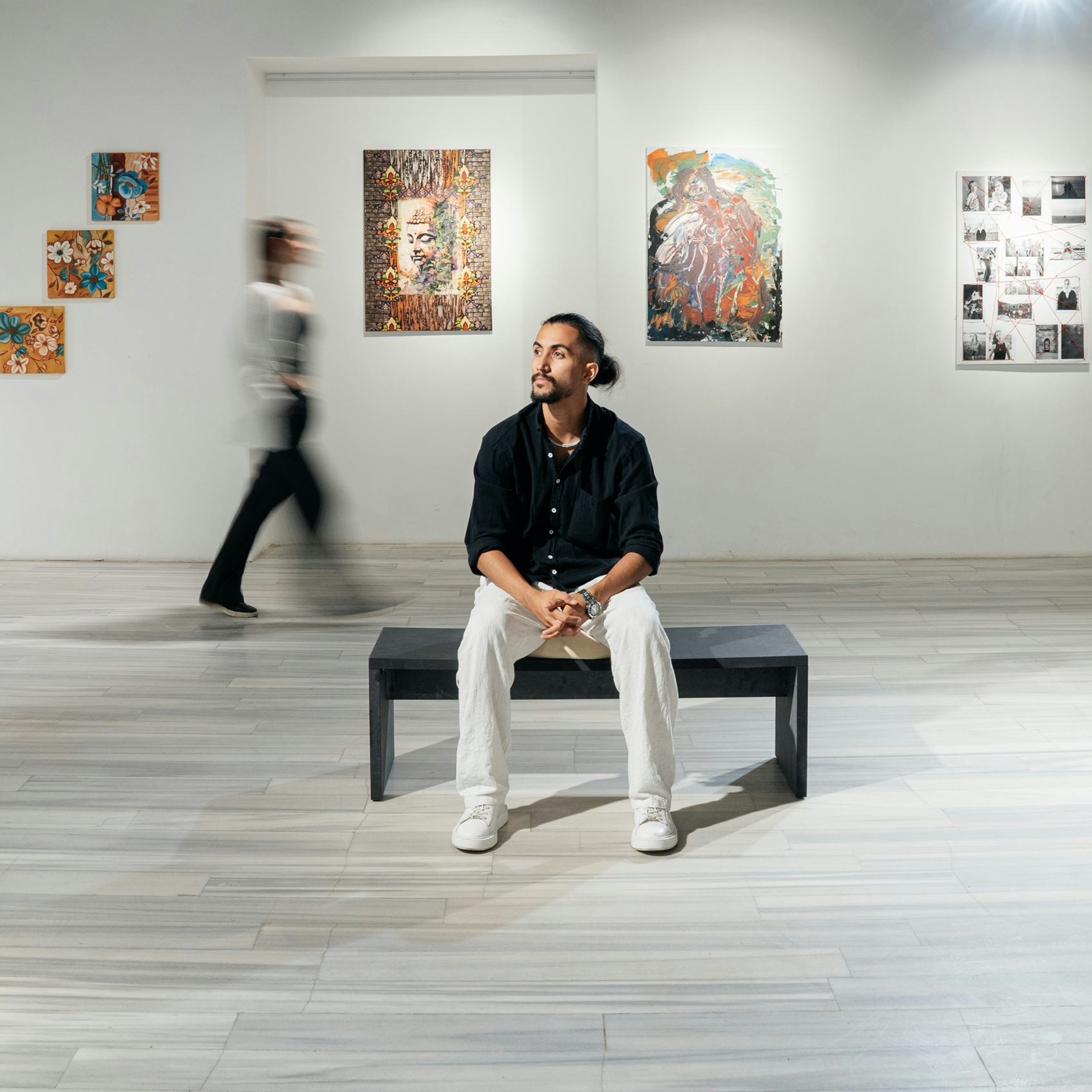 A person sits on a bench in a gallery, surrounded by colourful paintings and mixed-media works. Another visitor walks past, blurred in motion.